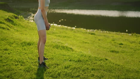 Close-up-from-the-side:-A-young-woman-sports-squats-with-dumbbells-in-the-morning-in-a-park-at-sunset-near-a-pond-or-river